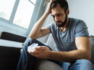 Depressed gloomy bearded man holding a pill organizer and looking at it while being ill