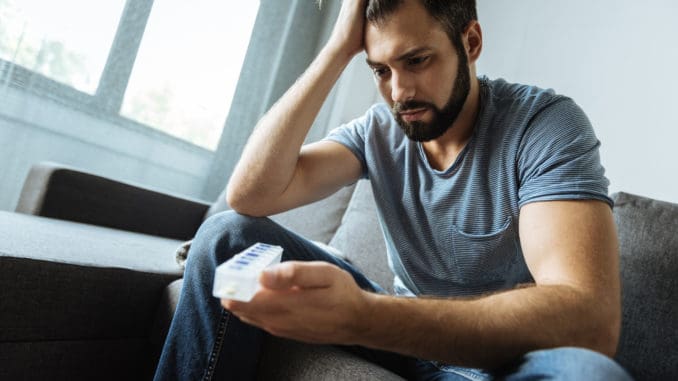 Depressed gloomy bearded man holding a pill organizer and looking at it while being ill