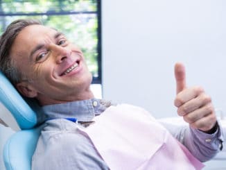 Portrait of smiling man showing thumbs up while sitting on chair at dentist clinic