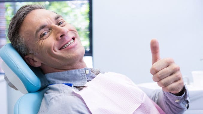 Portrait of smiling man showing thumbs up while sitting on chair at dentist clinic