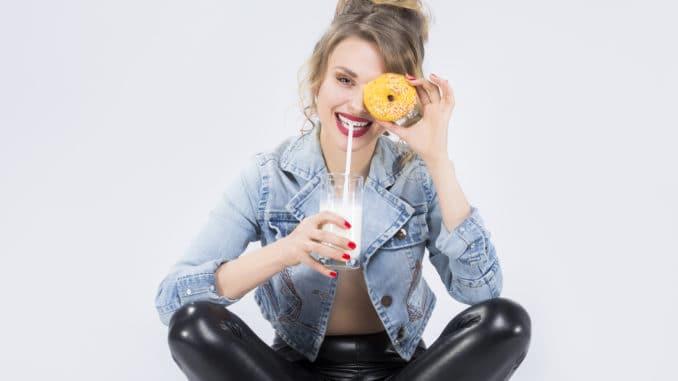 Food Ideas and Concepts. Young Caucasian Blond Girl Having Fun w Food Ideas and Concepts. Young Caucasian Blond Girl Having Fun with Cup of MIlk and Donut Bagel. Drinking Milk Through Straw