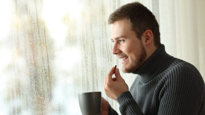 dreamstime_m_107730487 Side view portrait of a happy man taking a pill looking through a window in a rayny day of winter at home