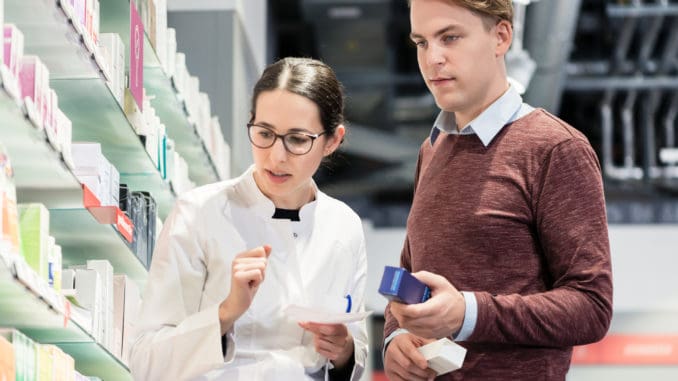 Low-angle view of a handsome young men holding two prescribed medicines while looking at various pharmaceutical products in a modern drugstore with helpful pharmacists