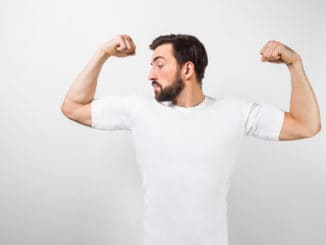 A handsome confident young man standing and showing big muscles on his hands. He is looking at one of them and very proud of them. He is wearing a white t-shirt, on white background