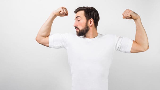 A handsome confident young man standing and showing big muscles on his hands. He is looking at one of them and very proud of them. He is wearing a white t-shirt, on white background