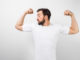 A handsome confident young man standing and showing big muscles on his hands. He is looking at one of them and very proud of them. He is wearing a white t-shirt, on white background