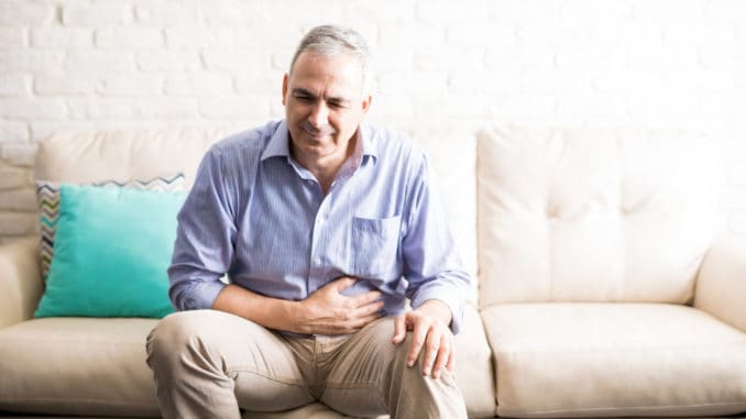dreamstime_m_110981845 Old man feeling unwell with a stomach ache while sitting on a sofa at home