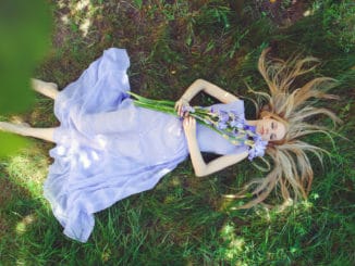 Attractive young girl with blonde hair and natural make-up smelling blue purple iris flowers lying on grass outdoors, tenderness and softness on nature background.