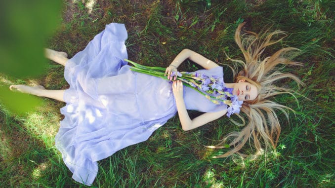 Attractive young girl with blonde hair and natural make-up smelling blue purple iris flowers lying on grass outdoors, tenderness and softness on nature background.