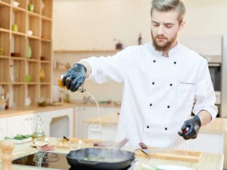 Portrait of handsome professional chef working in modern restaurant kitchen standing at wooden table and adding oils to delicious dishes, copy space