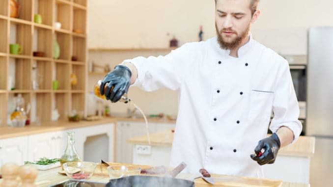 Portrait of handsome professional chef working in modern restaurant kitchen standing at wooden table and adding oils to delicious dishes, copy space