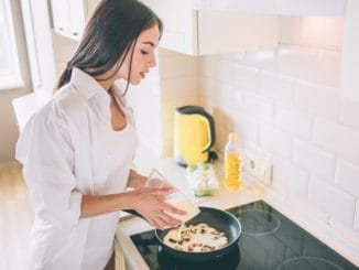 Beautiful young woman is cooking breakfast. She is pouring mixed eggs and milk into frying pan with mushrooms. Girl is cooking breakfast.