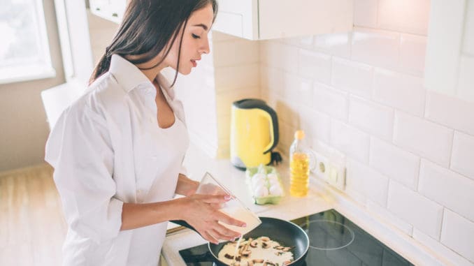 Beautiful young woman is cooking breakfast. She is pouring mixed eggs and milk into frying pan with mushrooms. Girl is cooking breakfast.