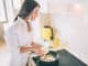 Beautiful young woman is cooking breakfast. She is pouring mixed eggs and milk into frying pan with mushrooms. Girl is cooking breakfast.