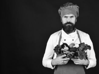 Man with beard on black background, copy space. Cook with serious face in burgundy uniform holds vegetables in wicker bowl. Chef holds lettuce, tomato, pepper and mushrooms. Vegetarian diet concept.