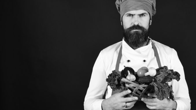 Man with beard on black background, copy space. Cook with serious face in burgundy uniform holds vegetables in wicker bowl. Chef holds lettuce, tomato, pepper and mushrooms. Vegetarian diet concept.