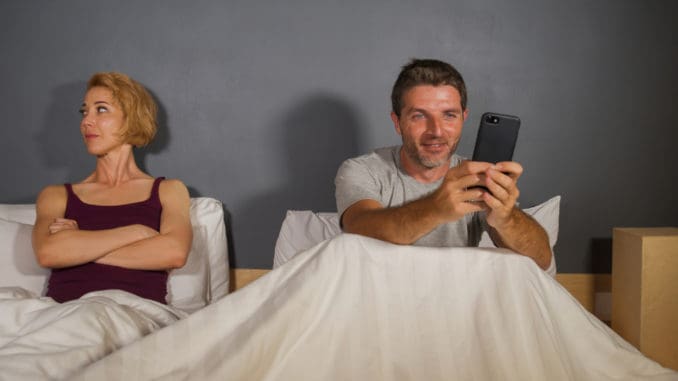 husband or boyfriend using mobile phone in bed and suspicious frustrated wife or girlfriend feeling upset suspecting betrayal and cheating in man woman couple relationship boyfriend using mobile phone in bed