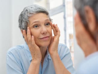 Beautiful senior woman checking her face skin and looking for blemishes. Portrait of mature woman massaging her face while checking wrinkled eyes in the mirror. Wrinkled lady with grey hair checking wrinkles around eyes, aging process.
