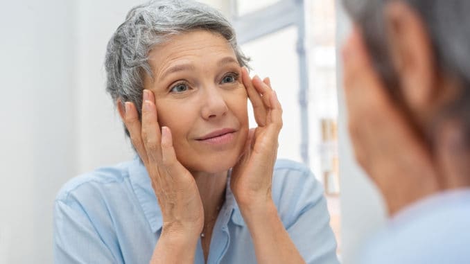 Beautiful senior woman checking her face skin and looking for blemishes. Portrait of mature woman massaging her face while checking wrinkled eyes in the mirror. Wrinkled lady with grey hair checking wrinkles around eyes, aging process.