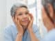 Beautiful senior woman checking her face skin and looking for blemishes. Portrait of mature woman massaging her face while checking wrinkled eyes in the mirror. Wrinkled lady with grey hair checking wrinkles around eyes, aging process.