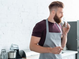handsome bearded young man in apron holding container with cinnamon sticks in kitchen