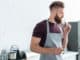 handsome bearded young man in apron holding container with cinnamon sticks in kitchen