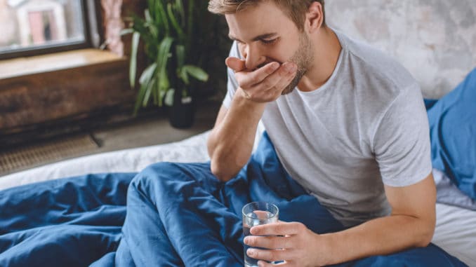 selective focus of young man taking pill and holding glass of water in bed at home young man taking pill