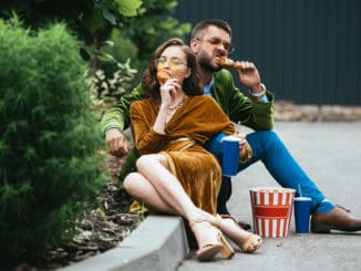 fashionable couple in velvet clothing eating fried chicken legs on street