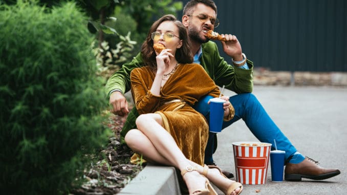 fashionable couple in velvet clothing eating fried chicken legs on street