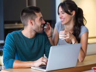 Shot of beautiful young women giving yogurt to her handsome boyfriend while they surfing in internet with laptop in the kitchen at home