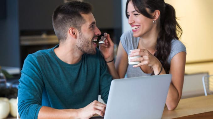 Shot of beautiful young women giving yogurt to her handsome boyfriend while they surfing in internet with laptop in the kitchen at home