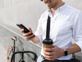 Photo of businesslike man 30s wearing sunglasses drinking takeaway coffee and using mobile phone while standing with bicycle along wall outdoor