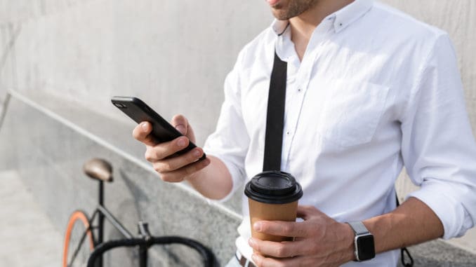 Photo of businesslike man 30s wearing sunglasses drinking takeaway coffee and using mobile phone while standing with bicycle along wall outdoor