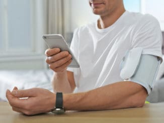 Man checking blood pressure with modern monitor and smartphone at table indoors, closeup. Cardiology concept