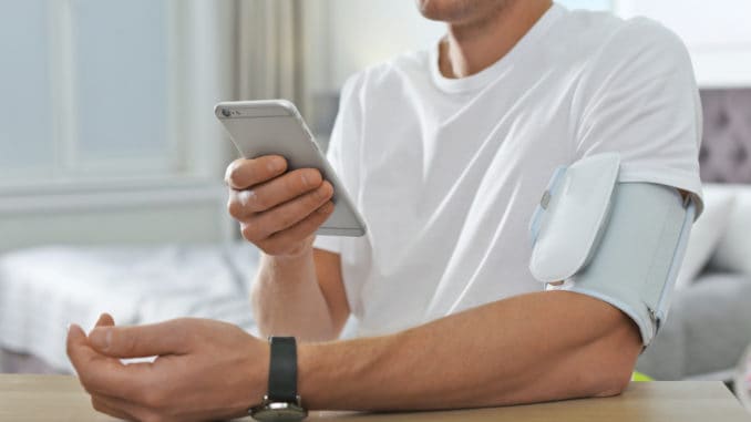 Man checking blood pressure with modern monitor and smartphone at table indoors, closeup. Cardiology concept
