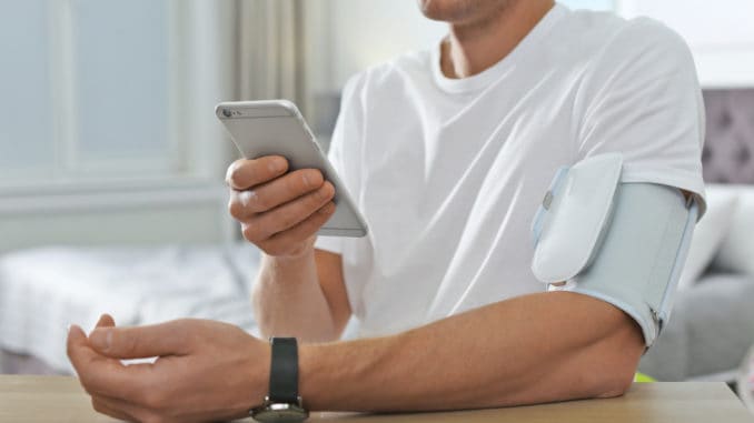 dreamstime_m_141599178 Man checking blood pressure with modern monitor and smartphone at table indoors, closeup. Cardiology concept