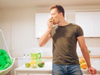 Young smiling handsome man bites organic green apple while cooking fresh fruits in the kitchen. Healthy eating. Vegetarian meal. Diet detox