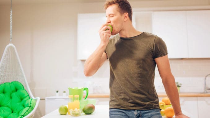 Young smiling handsome man bites organic green apple while cooking fresh fruits in the kitchen. Healthy eating. Vegetarian meal. Diet detox