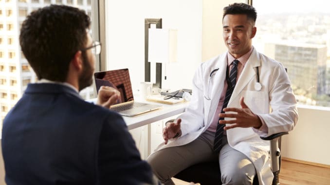 Over The Shoulder View Of Man Having Consultation With Male Doctor In Hospital Office Man Having Consultation With Male Doctor In Hospital Office