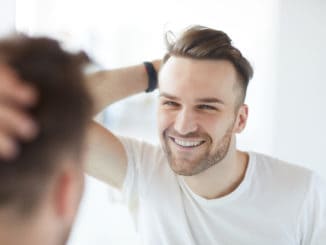 Portrait of handsome young man with lush hair and short stubble looking at his reflection in mirror and smiling, copy space