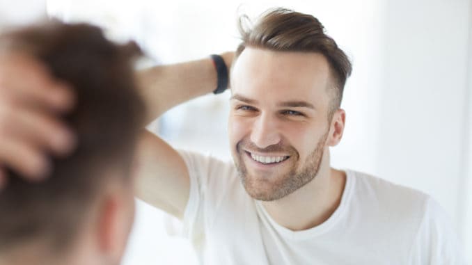 Portrait of handsome young man with lush hair and short stubble looking at his reflection in mirror and smiling, copy space