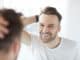 Portrait of handsome young man with lush hair and short stubble looking at his reflection in mirror and smiling, copy space