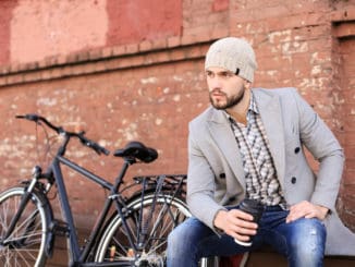 Handsome young man in grey coat and hat sitting on a bench relaxed drinking coffee and thinking near his bicycle.