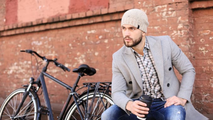 Handsome young man in grey coat and hat sitting on a bench relaxed drinking coffee and thinking near his bicycle.