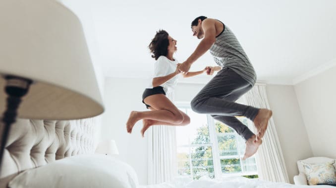 Man and women holding hands and jumping together on bed.