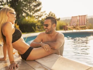 Couple in love at a poolside summer party, sitting at the edge of a swimming pool, sunbathing, drinking cocktails and having fun