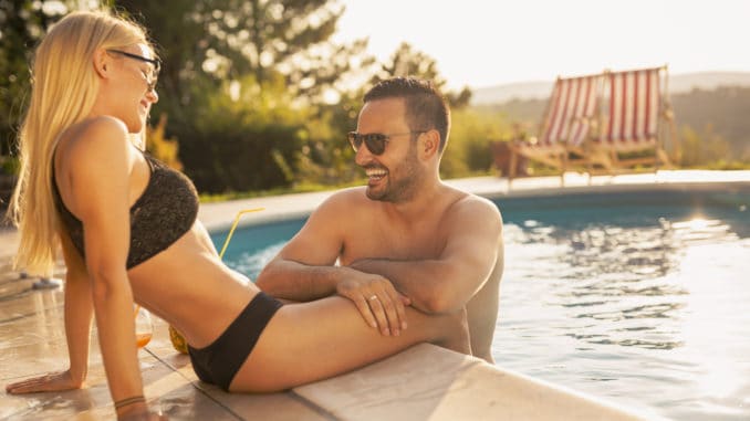 Couple in love at a poolside summer party, sitting at the edge of a swimming pool, sunbathing, drinking cocktails and having fun