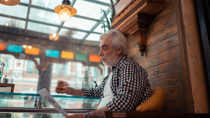 Grey-haired man wearing checked shirt reading news and drinking alcohol