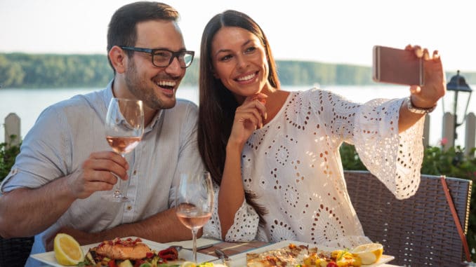 Smiling happy young couple posing for a selfie, eating dinner in a riverside restaurant. Husband and wife celebrating anniversary, having fish dinner in outdoor restaurant