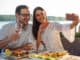 Smiling happy young couple posing for a selfie, eating dinner in a riverside restaurant. Husband and wife celebrating anniversary, having fish dinner in outdoor restaurant